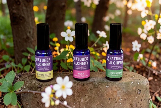 Three Natures Alchemy hydrating facial mists in Lemon Balm, Rose and Spearmint, displayed on a mossy stone surrounded by spring wildflowers