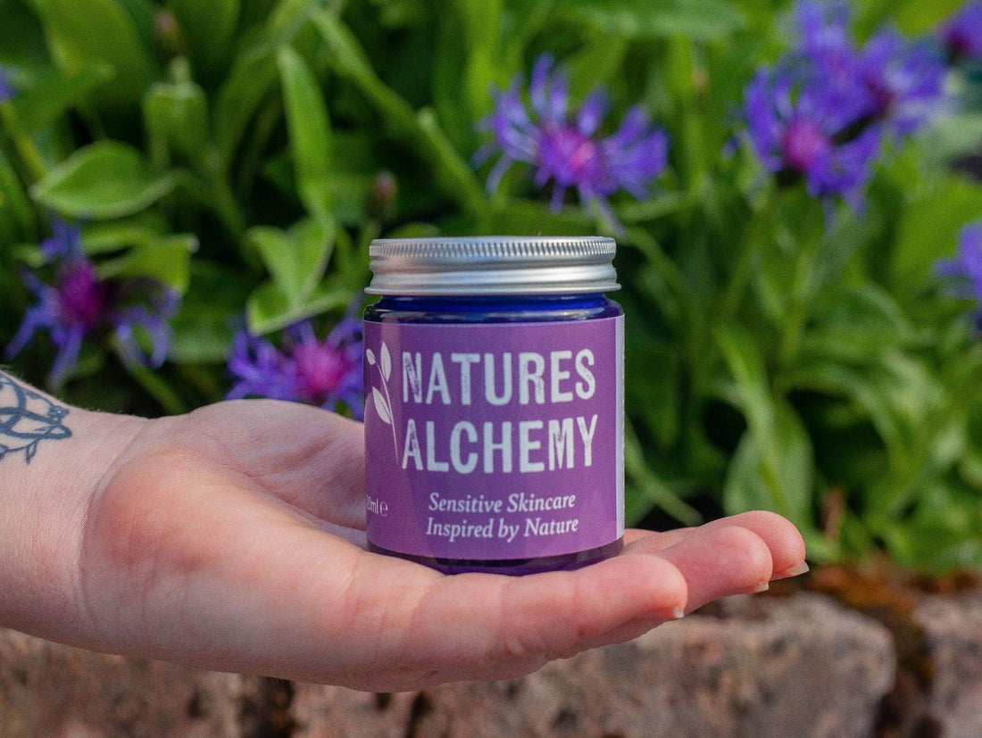 Hand holding a Natures Alchemy solid body oil jar with purple wildflowers in the background, natural Irish skincare brand
