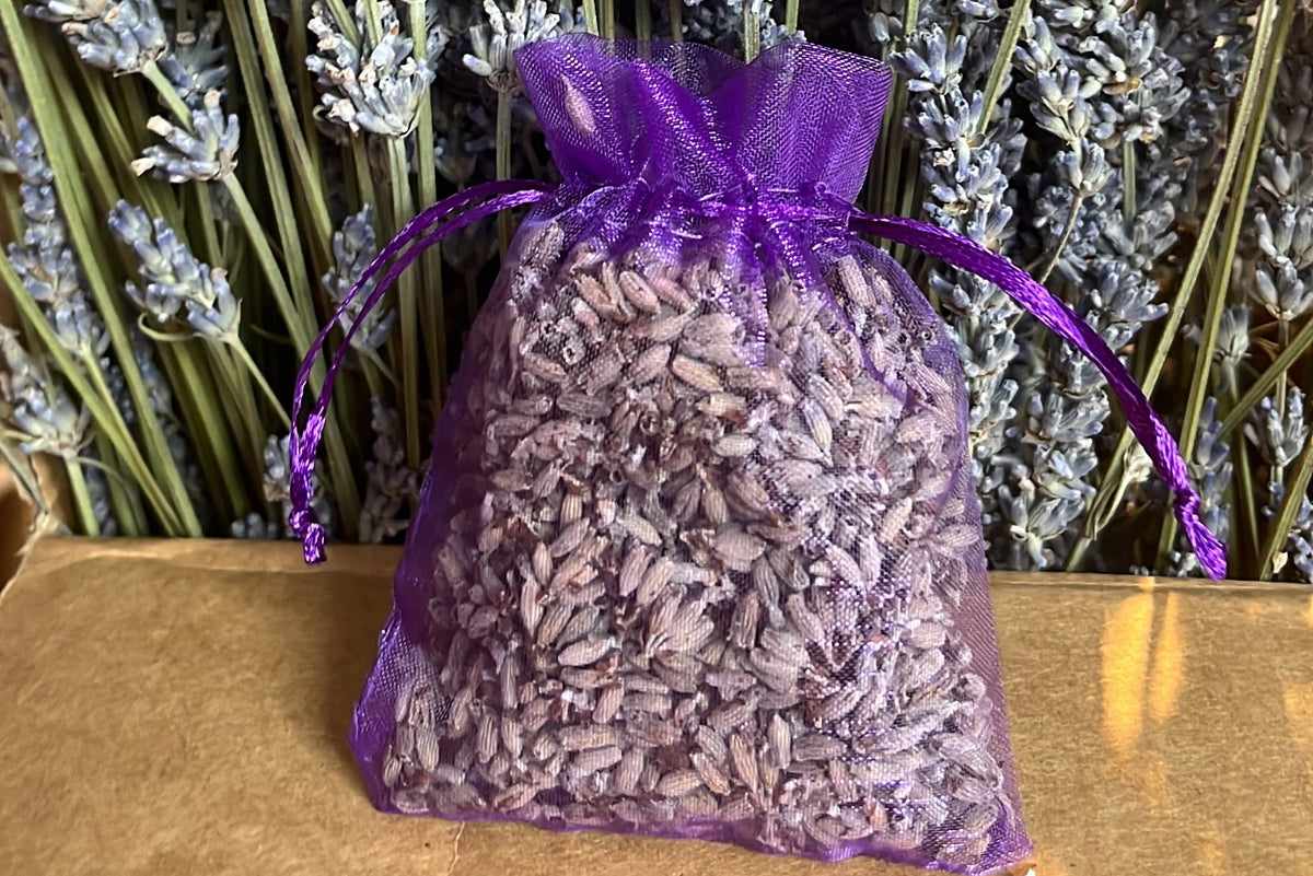A small purple sheer drawstring pouch filled with dried lavender buds, placed on a tan surface in front of a dense background of lavender stems.